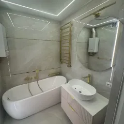 Nowoczesna łazienka w odcieniach szarości z białymi elementami, złote akcenty w armaturze i oświetleniu, wanna wolnostojąca, okrągłe lustro z podświetleniem, podwieszana szafka umywalkowa...