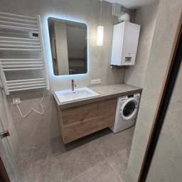 GLAZURNIK Rafał Matura - Nowoczesna łazienka z szarymi płytkami, drewnianą szafką pod umywalką, miedzianą baterią, podświetlanym lustrem, pralką, białym piecem gazowym i grzejnikiem drabinkowym.