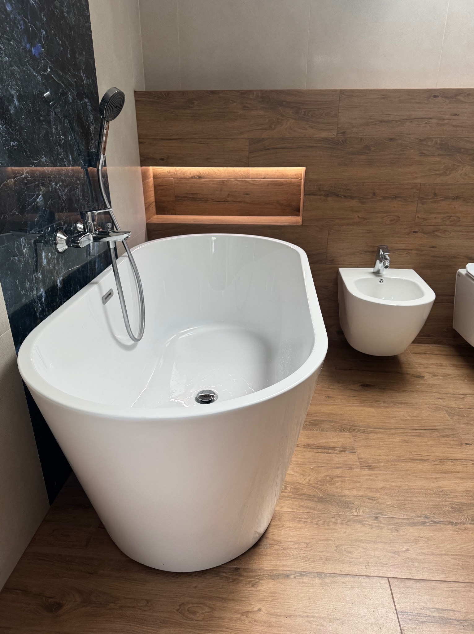 Nowoczesna łazienka z wolnostojącą białą wanną, bidetem i drewnopodobnymi płytkami na podłodze i ścianie, z oświetloną wnęką.