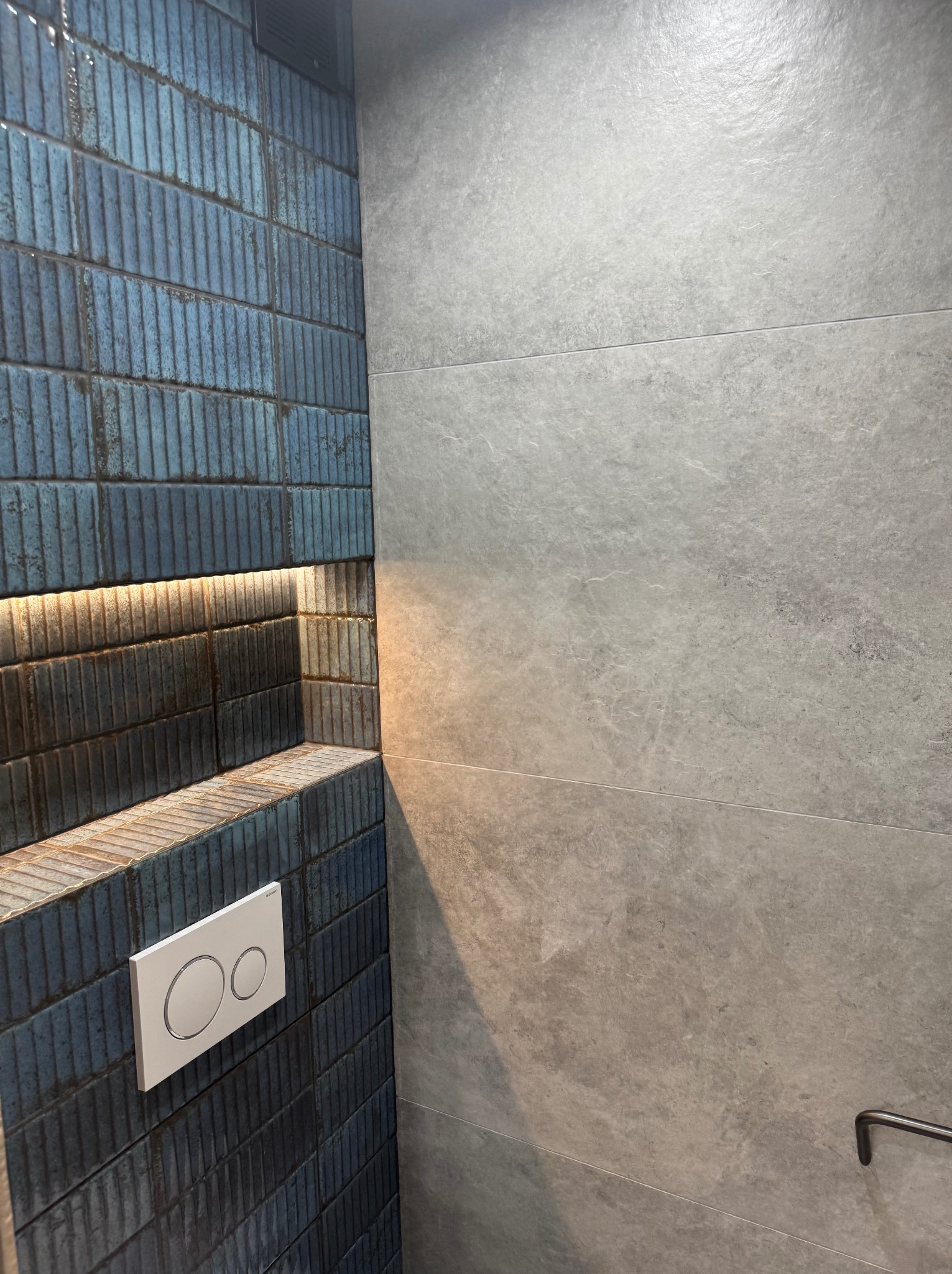 Niebieska, teksturowana ściana z płytek w łazience, z podświetlaną wnęką i przyciskiem spłuczki, kontrastująca z gładką, szarą ścianą z płytek.