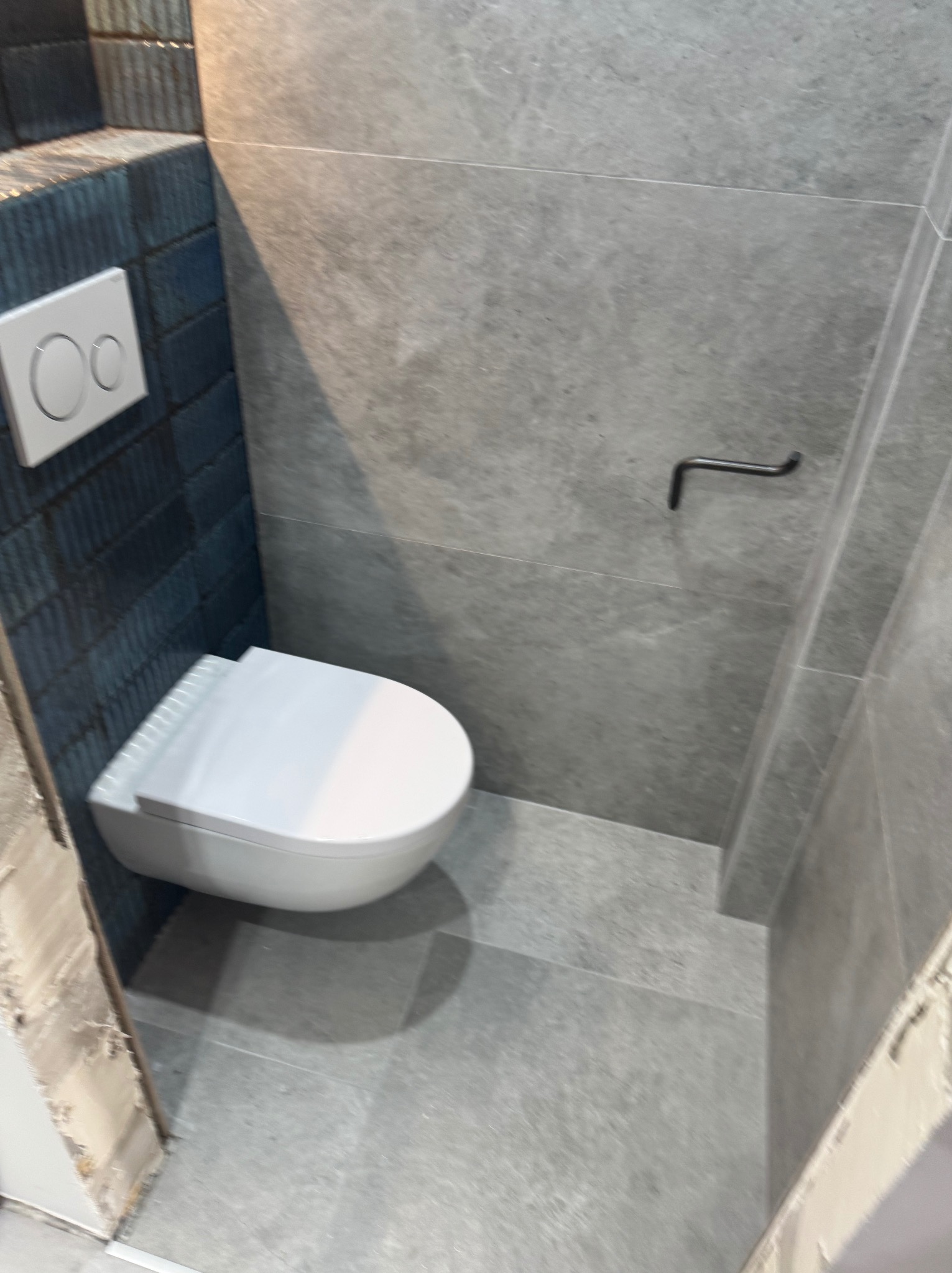 Wisząca toaleta z przyciskiem spłukującym na tle ściany z ciemnej, strukturalnej glazury i szarej, gładkiej płytki.