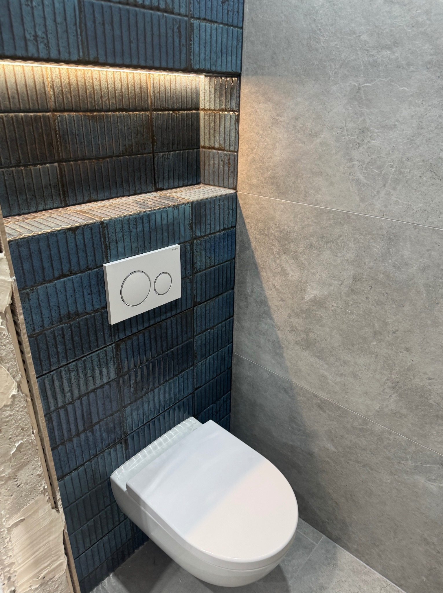 Nowoczesna łazienka po remoncie: wisząca toaleta z przyciskiem Geberit na ścianie z ciemnoniebieskich, pionowo ułożonych płytek, podświetlona półka, szara ściana wykończona płytami imitującymi kamień.
