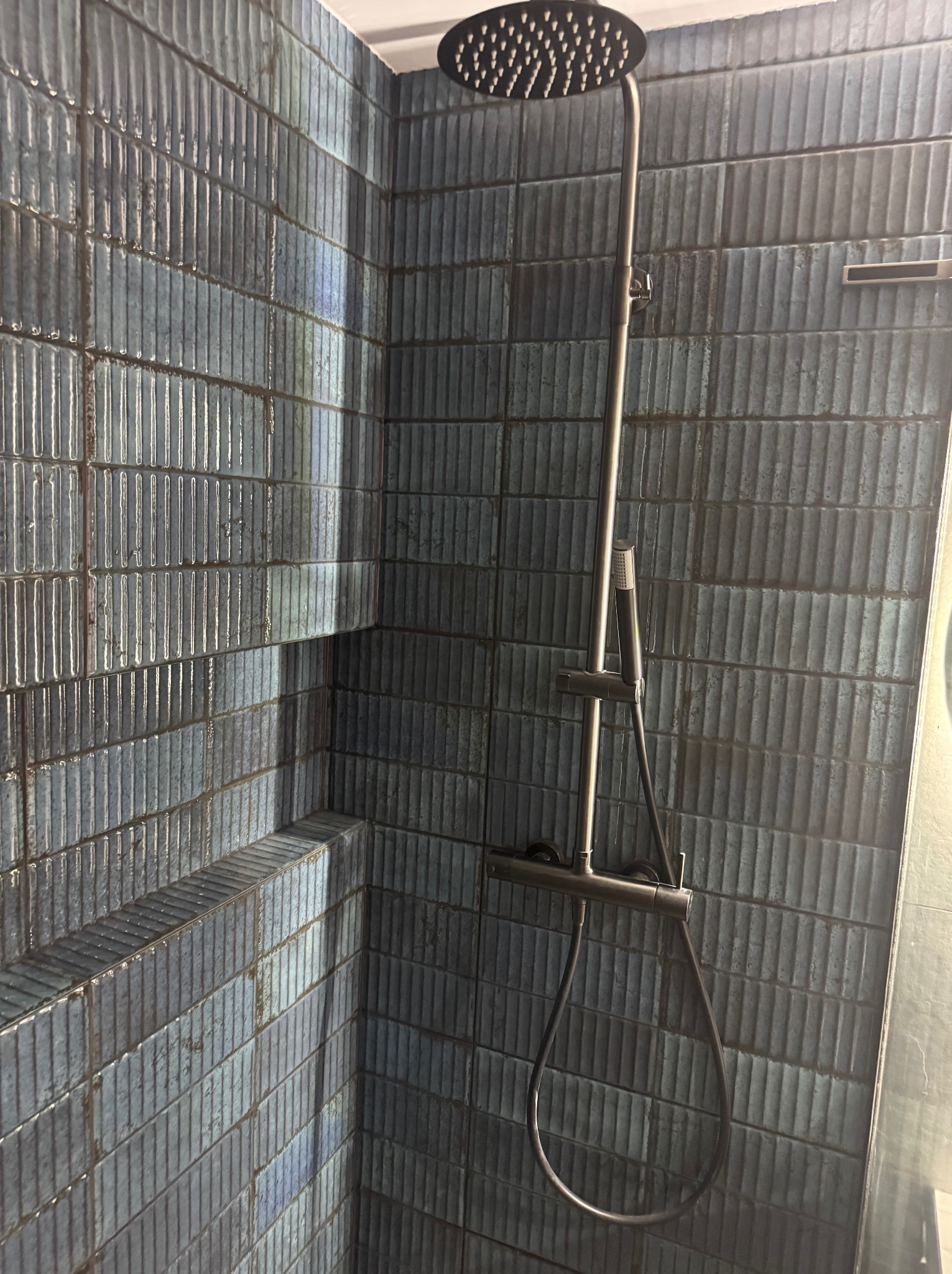 Kabina prysznicowa z ciemnymi, pionowo ułożonymi płytkami imitującymi cegłę, deszczownica i bateria w kolorze chromu.