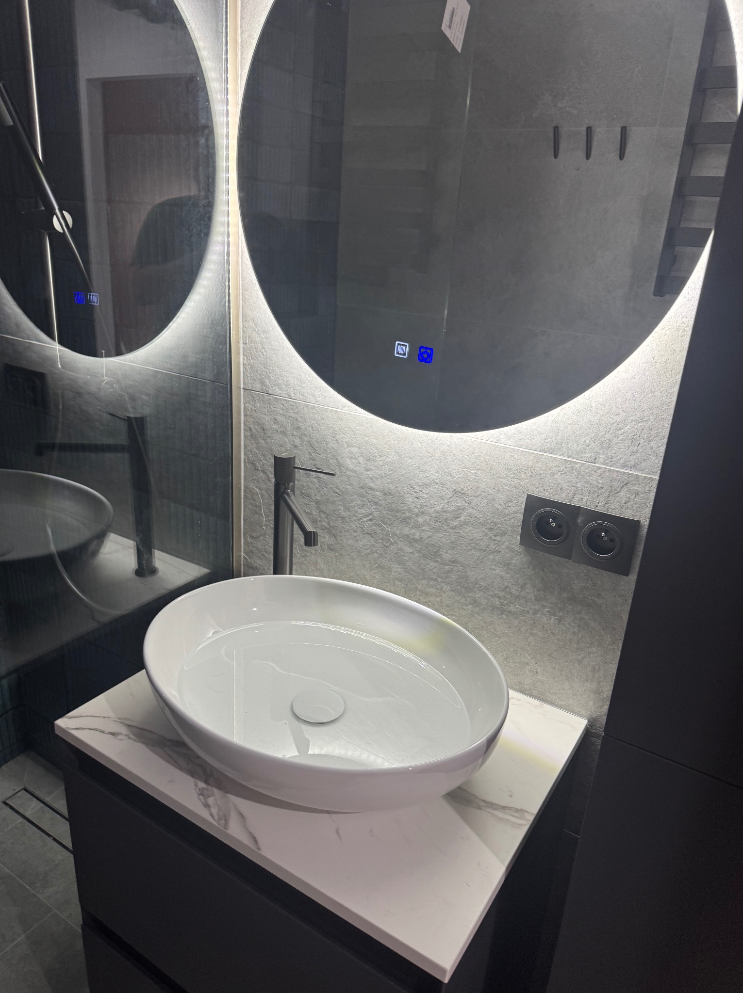 Nowoczesna łazienka z okrągłym lustrem podświetlanym LED, białą umywalką nablatową na blacie imitującym marmur oraz szarą płytką na ścianie.