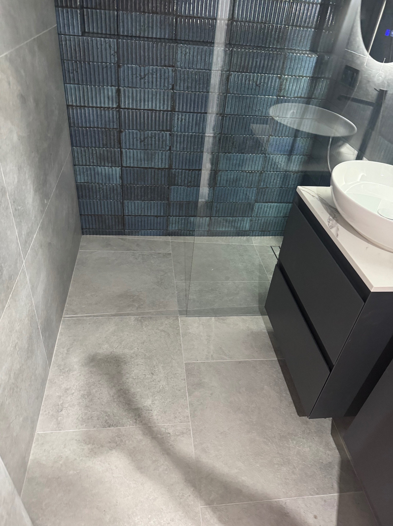 Nowoczesna łazienka z szarymi płytkami podłogowymi i ściennymi, akcentem niebieskich płytek w kabinie prysznicowej oraz czarną szafką z umywalką nablatową.