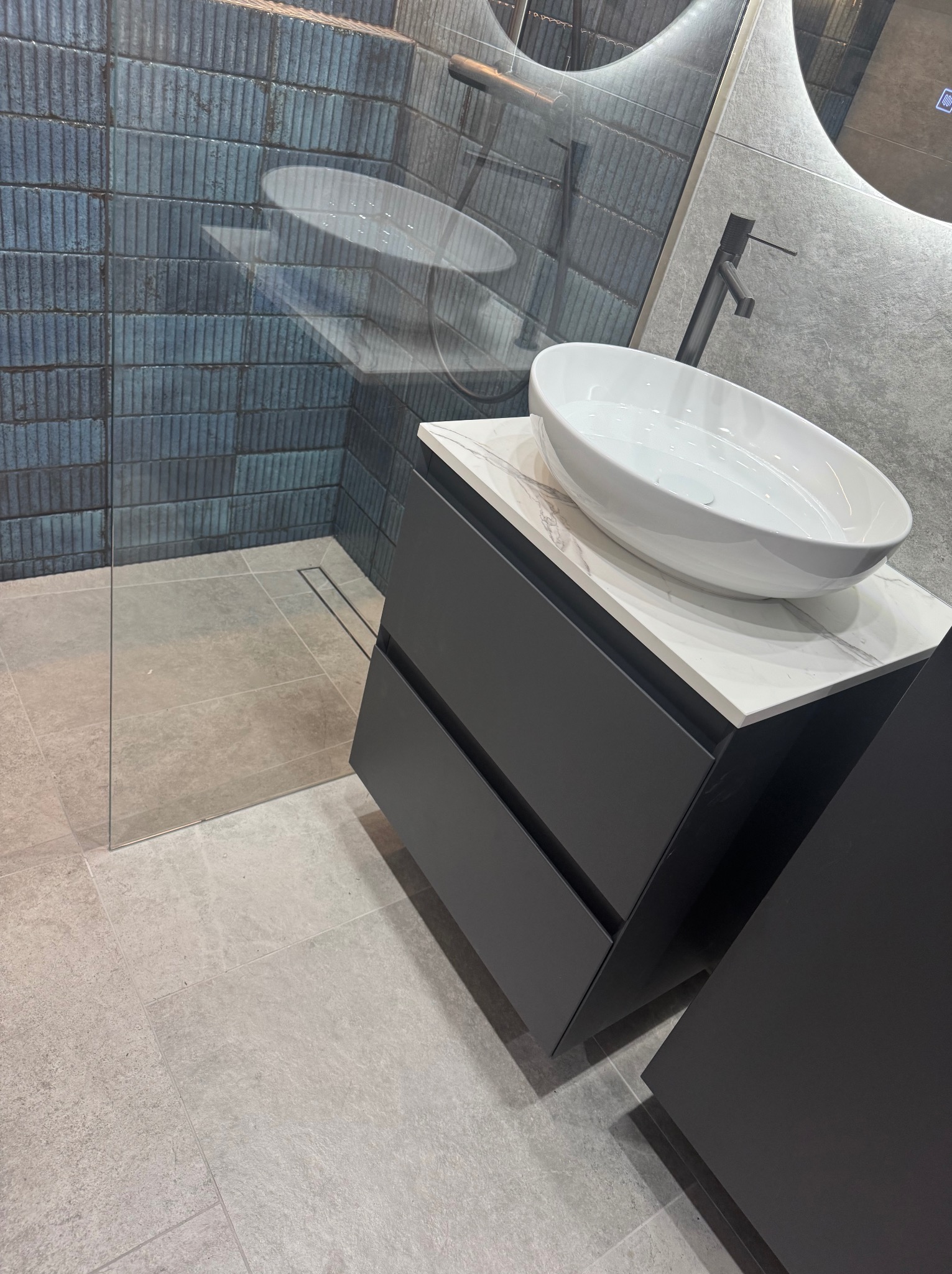 Nowoczesna łazienka z owalną umywalką nablatową, ciemnoszarą szafką z dwiema szufladami, kabiną prysznicową typu walk-in z niebieskimi płytkami i odpływem liniowym w podłodze.