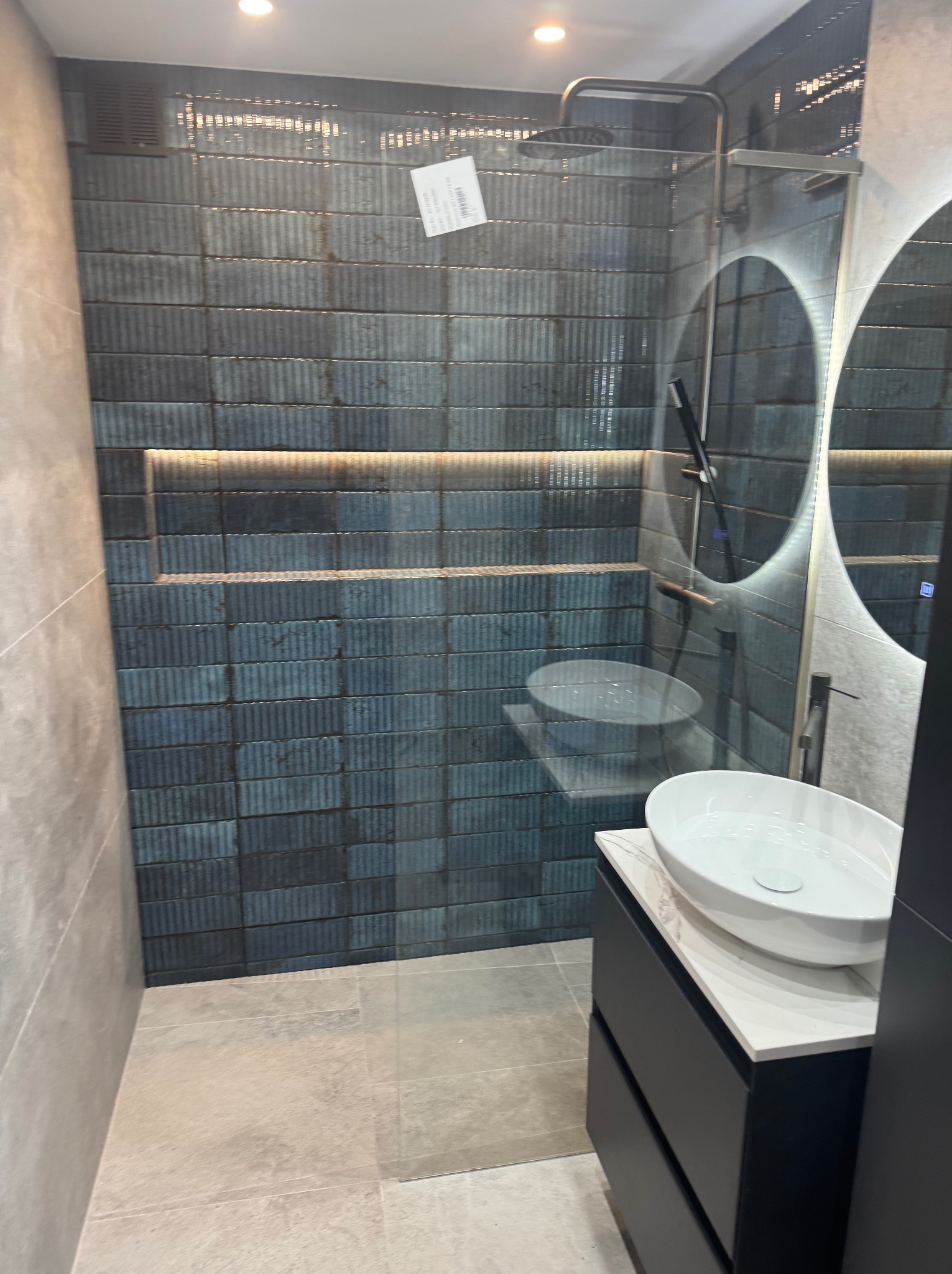 Nowoczesna łazienka z kabiną prysznicową typu walk-in wyłożoną ciemnymi, strukturalnymi płytkami, podświetloną wnęką, okrągłym lustrem z podświetleniem LED i białą umywalką nablatową na czarnej...