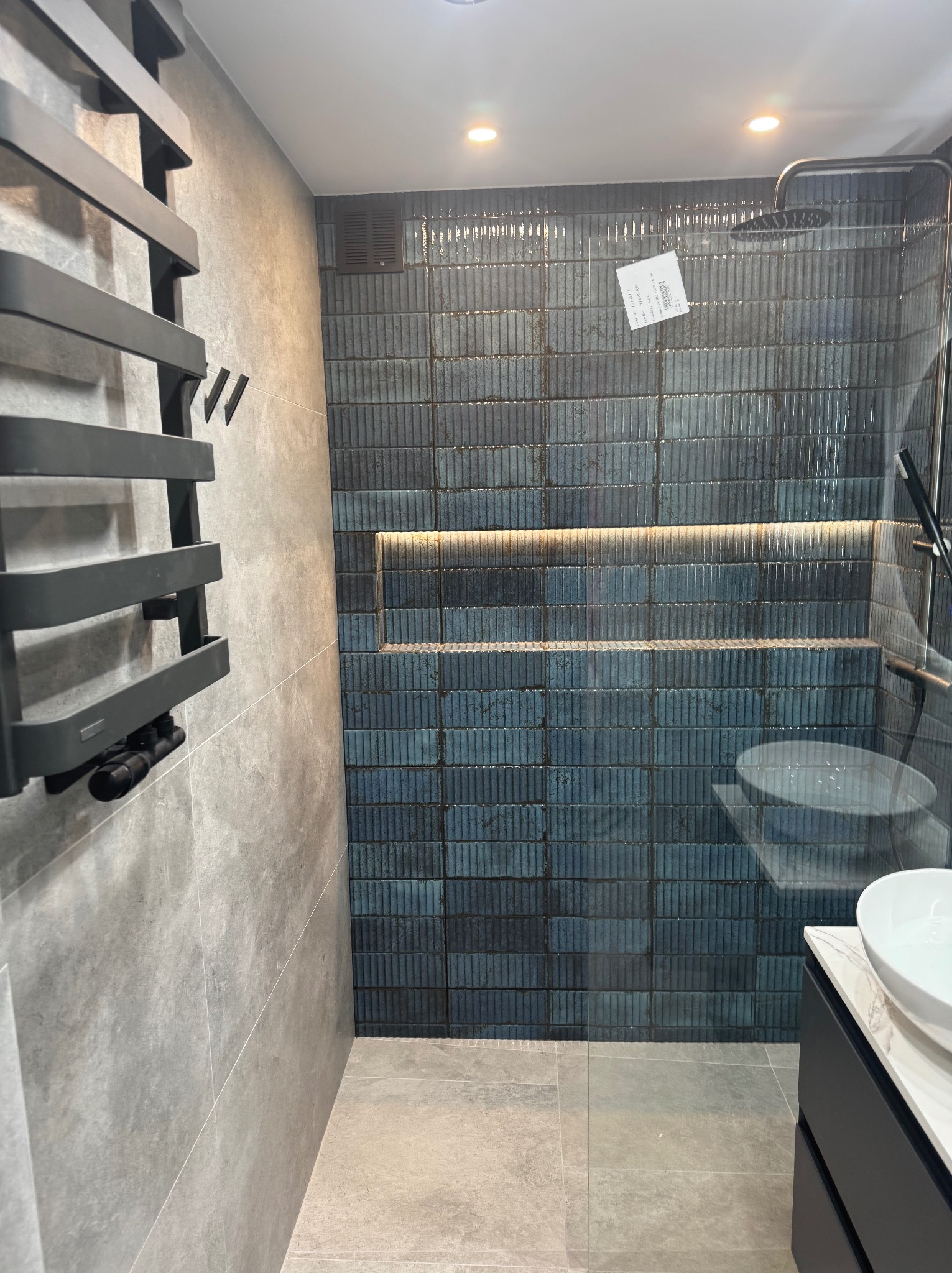 Nowoczesna łazienka z kabiną prysznicową wyłożoną pionowymi, niebieskimi płytkami, podświetloną wnęką, grzejnikiem drabinkowym i umywalką nablatową z szafką.