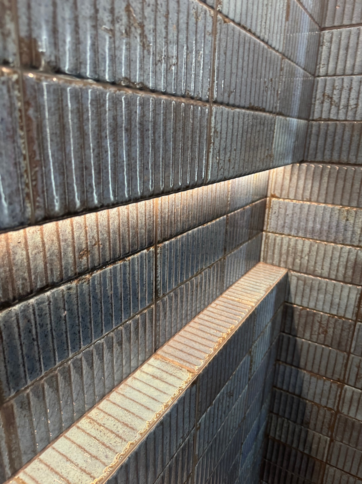 Fragment łazienki z szarymi, pionowo żłobionymi płytkami, podświetlonymi ciepłym światłem LED, tworzącymi półkę w rogu.