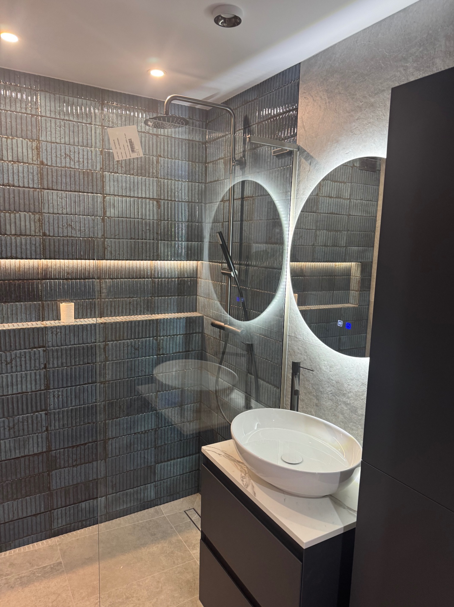 Nowoczesna łazienka z kabiną prysznicową typu walk-in, umywalką nablatową na szafce z blatem imitującym marmur, podświetlanym lustrem i płytkami w odcieniach szarości i granatu z fakturą cegiełek.