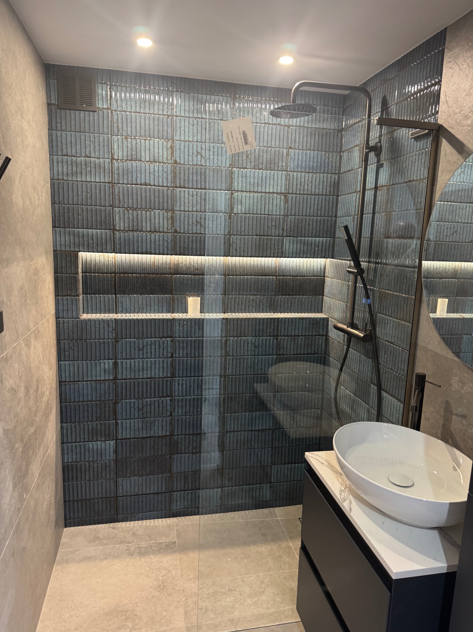 Nowoczesna kabina prysznicowa z ciemnymi, strukturalnymi płytkami, podświetlana wnęka, biała umywalka nablatowa na czarnej szafce z marmurowym blatem.