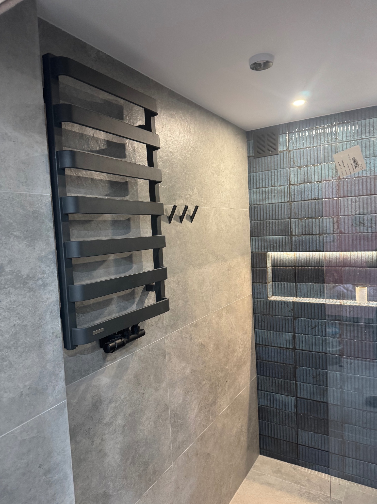 Nowoczesna łazienka z grafitowym grzejnikiem drabinkowym, szarą płytką imitującą kamień i kabiną prysznicową z niebieską mozaiką w pionowe paski oraz wnęką z podświetleniem LED.