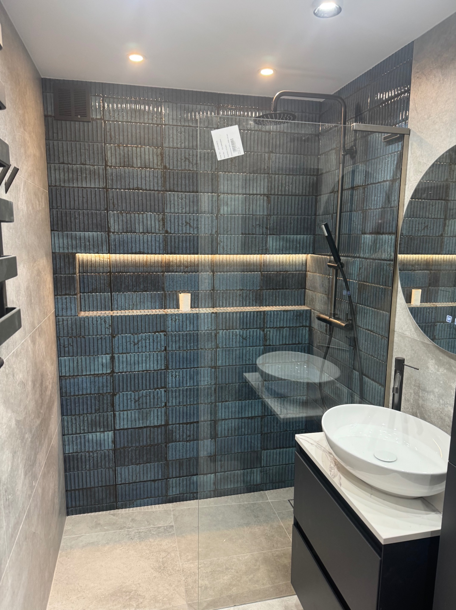 Nowoczesna łazienka z kabiną prysznicową typu walk-in, umywalką nablatową na czarnej szafce z blatem imitującym marmur oraz ścianą wyłożoną pionowymi, niebiesko-czarnymi płytkami z podświetlaną wnęką.