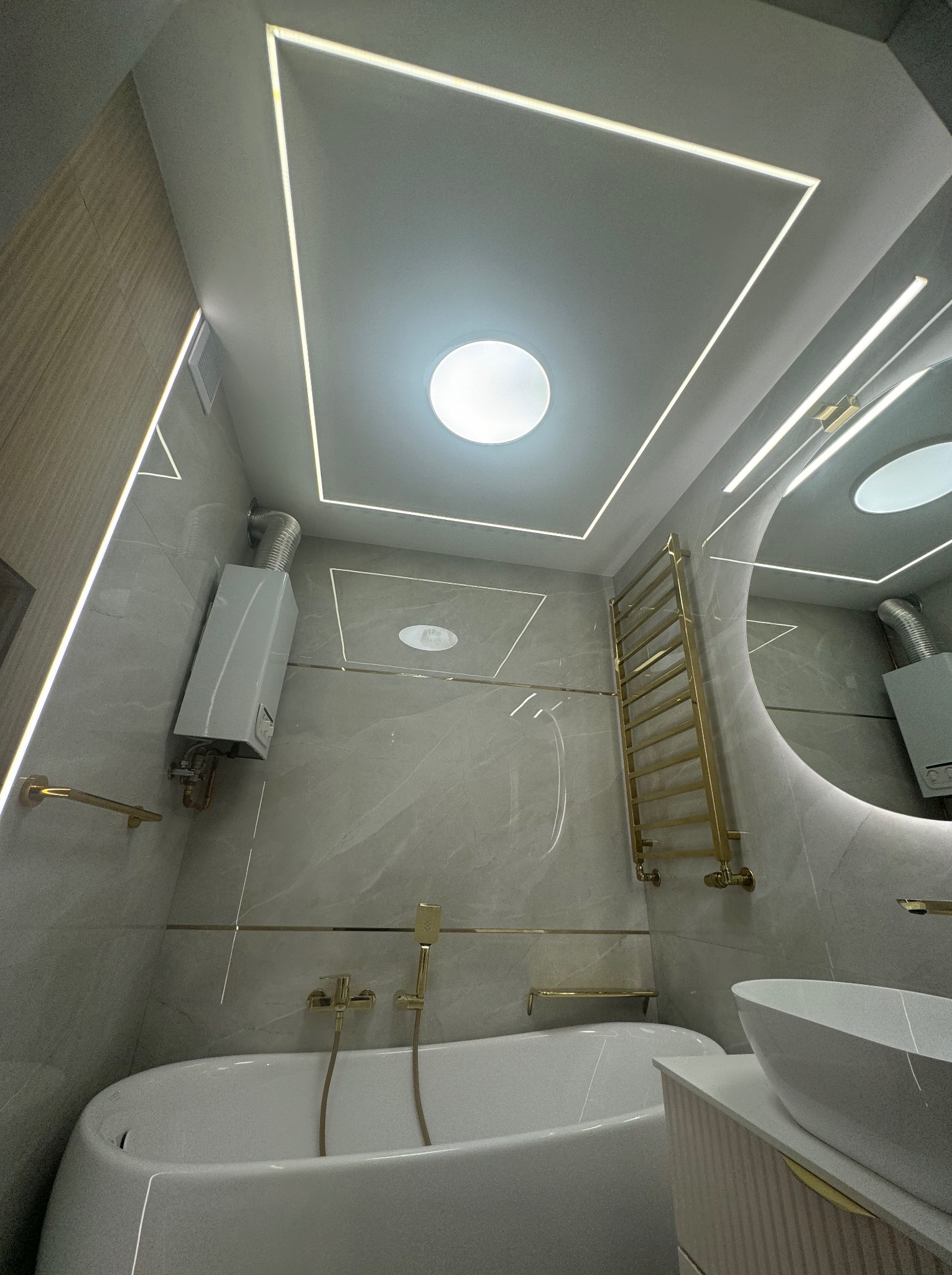 Elegancka łazienka z wanną, owalną umywalką, grzejnikiem drabinkowym w kolorze złota i podświetlanym sufitem z centralnym okrągłym świetlikiem.