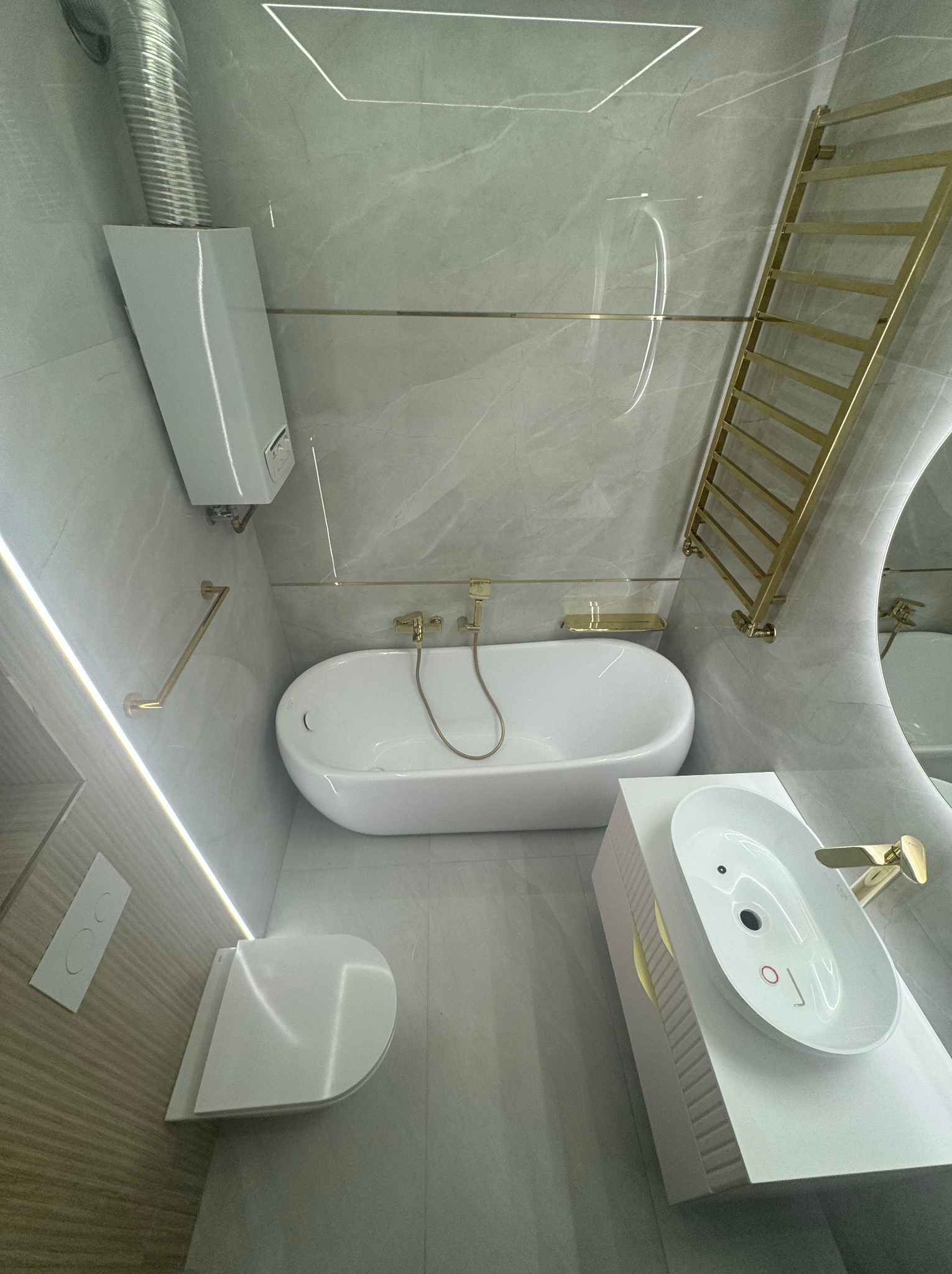 Nowoczesna łazienka z wanną wolnostojącą, umywalką nablatową na szafce z frezowanymi bokami, złotą armaturą i grzejnikiem drabinkowym, oświetleniem LED oraz piecem gazowym z odprowadzeniem spalin.