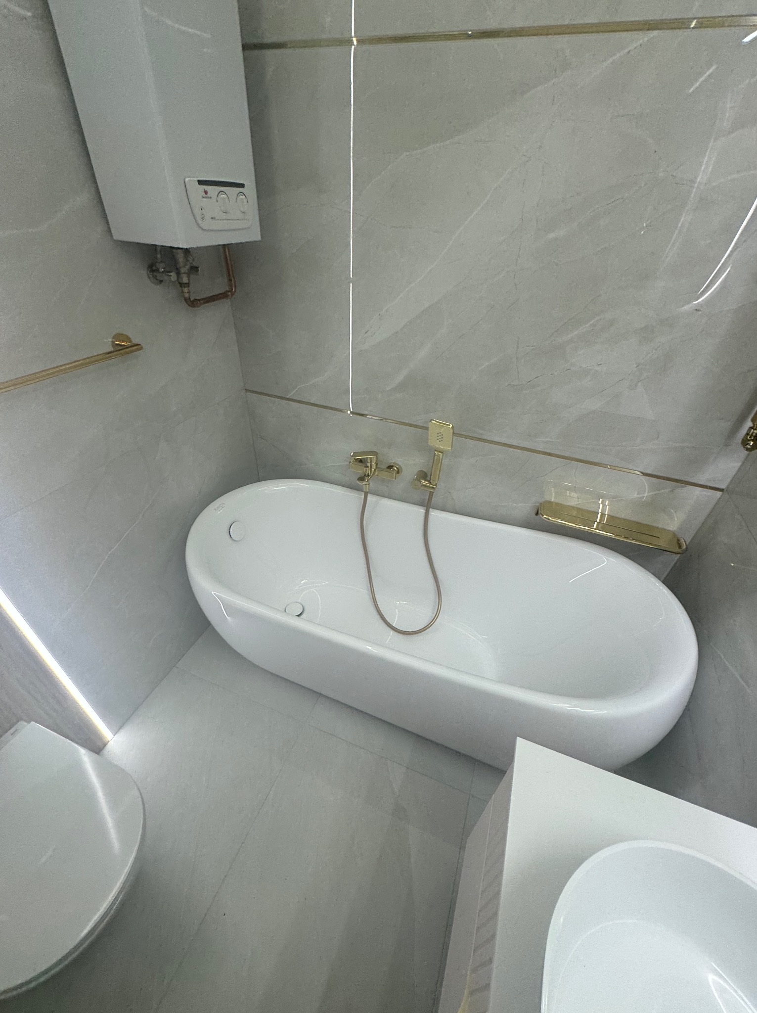 Nowoczesna łazienka w odcieniach szarości z białą wolnostojącą wanną, złotą armaturą i podświetleniem LED.