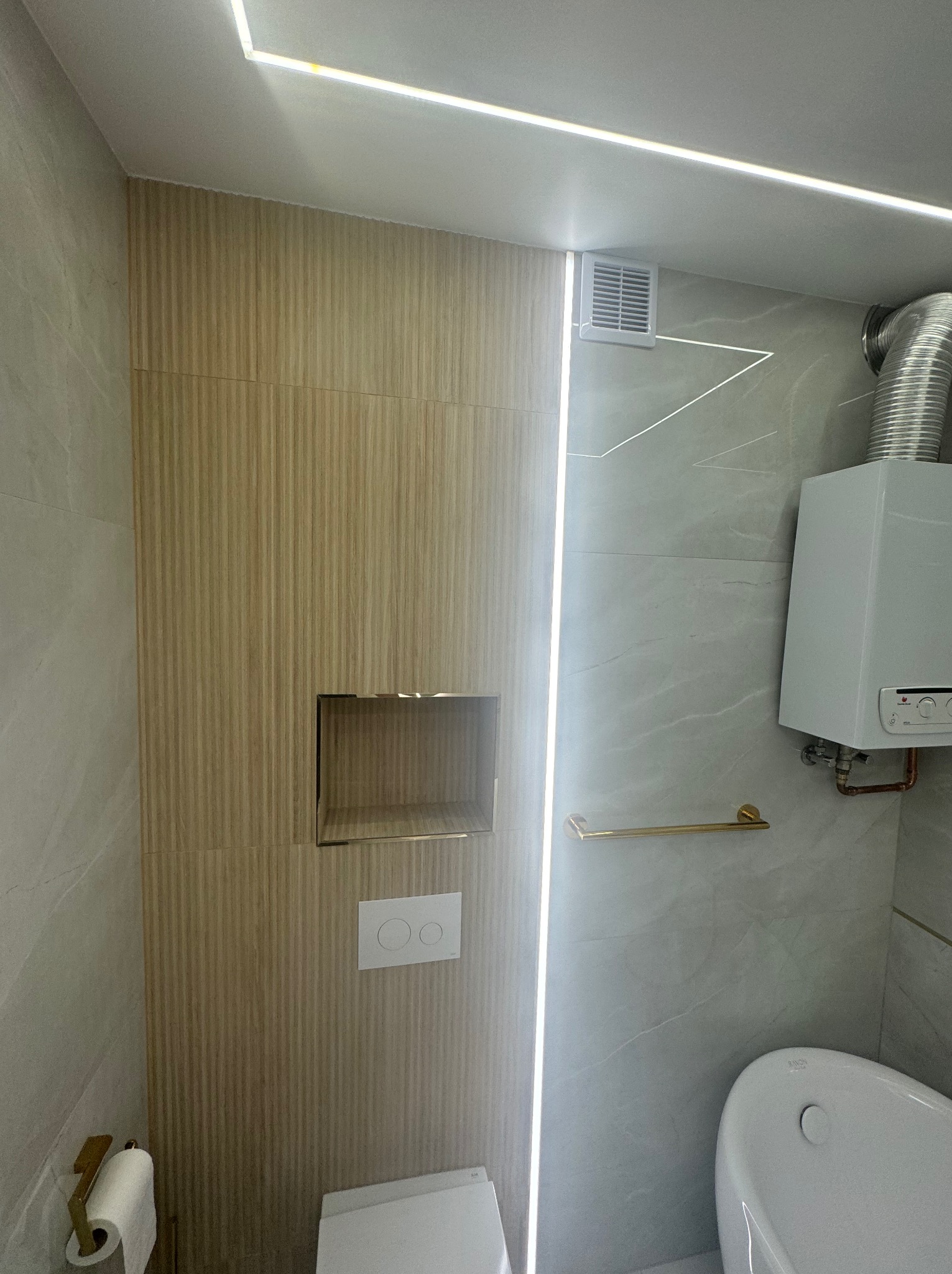 Nowoczesna łazienka z podświetlanym lustrem w niszy, wiszącą toaletą z białym panelem sterującym i grzejnikiem gazowym z chromowaną rurą odprowadzającą spaliny.