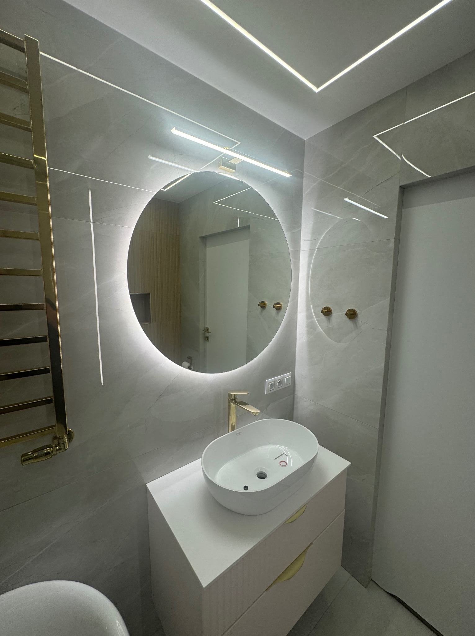 Nowoczesna łazienka z okrągłym lustrem podświetlanym LED, umywalką nablatową na białej szafce z złotymi uchwytami, grzejnikiem drabinkowym w kolorze złotym i oświetleniem sufitowym LED w kształcie...