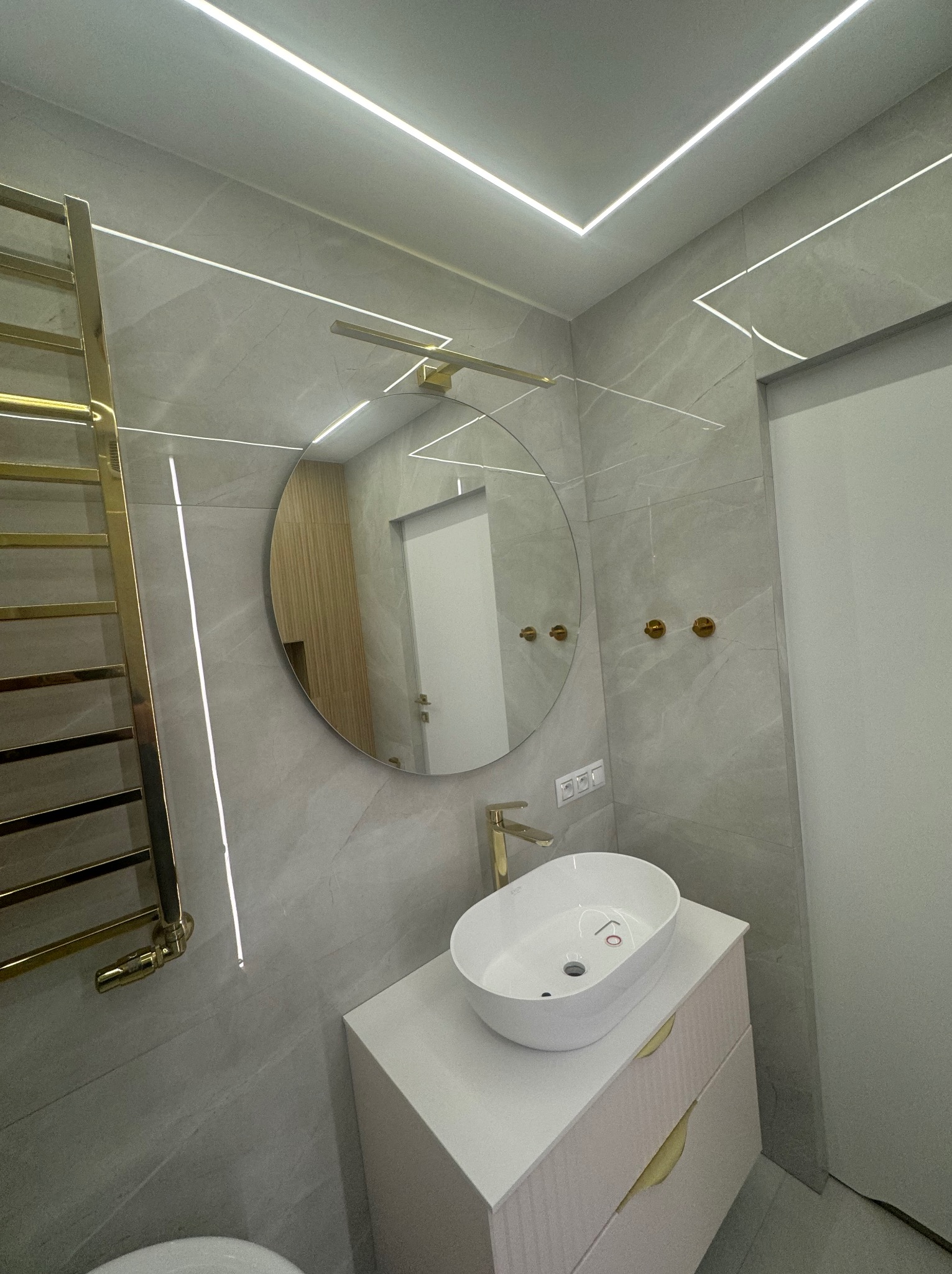 Nowoczesna łazienka z szarymi płytkami imitującymi marmur, okrągłym lustrem, białą umywalką nablatową, złotą armaturą i grzejnikiem drabinkowym oraz oświetleniem LED.