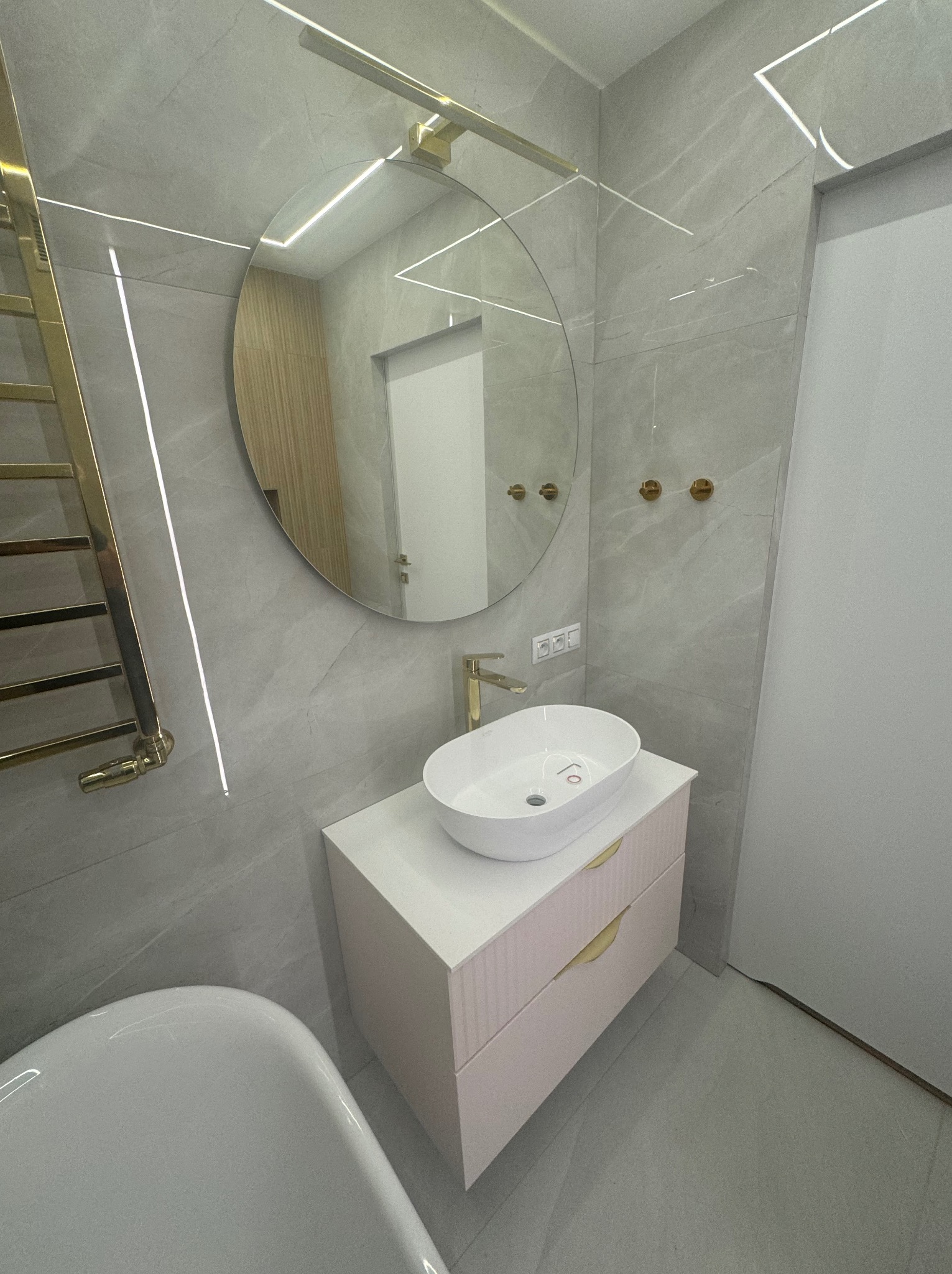 Nowoczesna łazienka z owalną umywalką nablatową, armaturą w kolorze złota, lustrem w okrągłej ramie i grzejnikiem drabinkowym, podświetlona ledami w płytkach imitujących marmur.