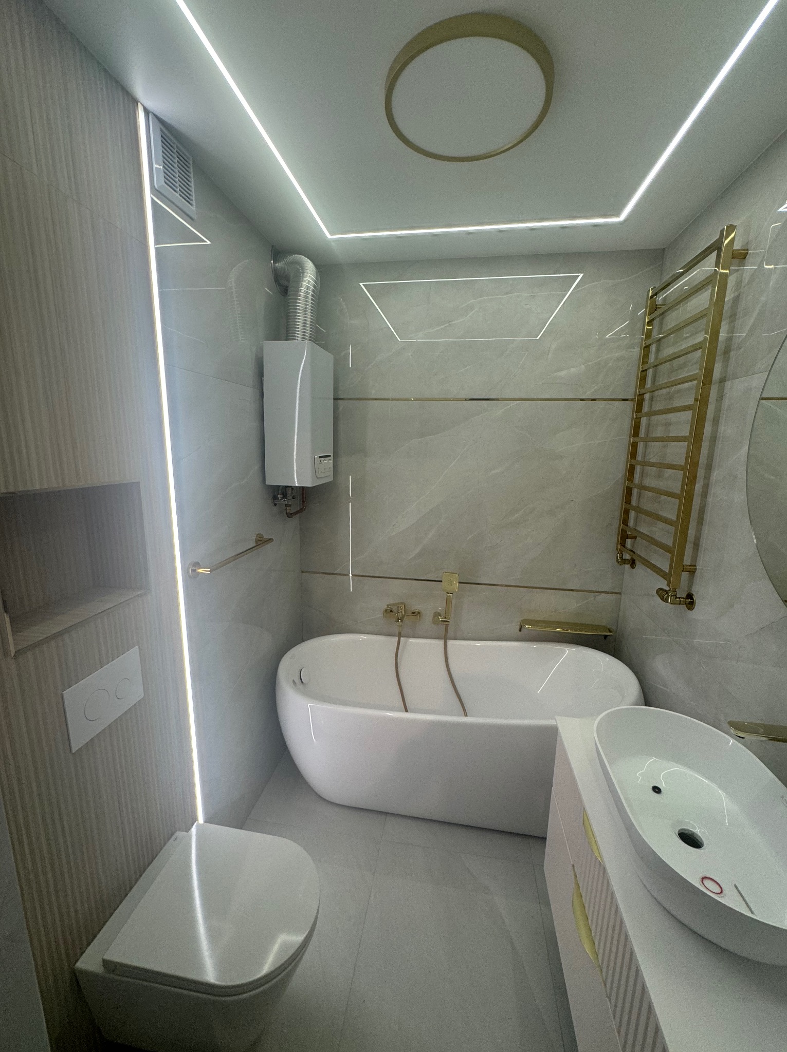 Nowoczesna łazienka z owalną wanną, wiszącą toaletą, umywalką nablatową i złotymi akcentami, oświetlona liniowymi lampami LED i z zamontowanym piecykiem gazowym.