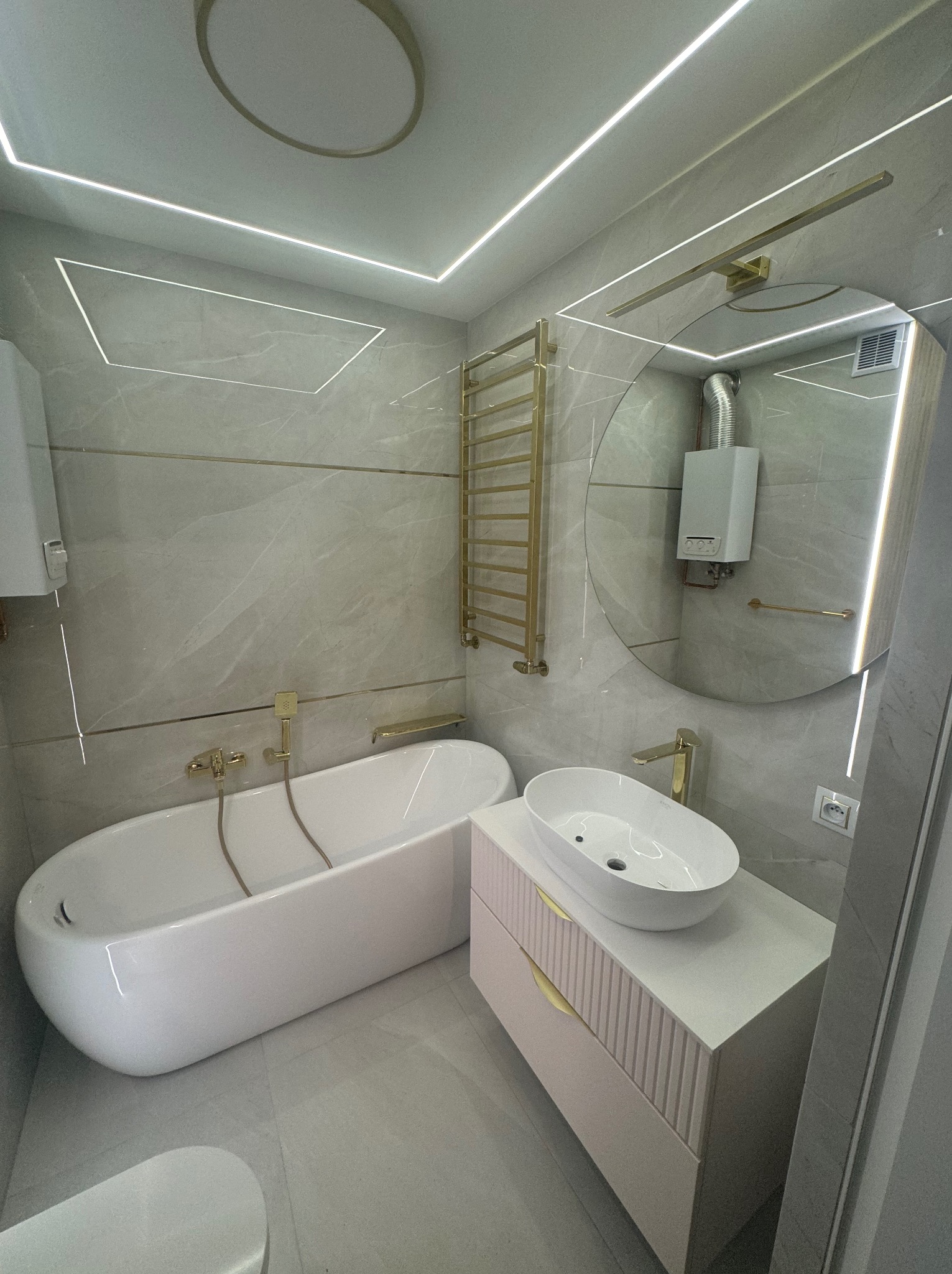 Nowoczesna łazienka w odcieniach szarości z białymi elementami, złote akcenty w armaturze i oświetleniu, wanna wolnostojąca, okrągłe lustro z podświetleniem, podwieszana szafka umywalkowa...