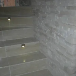 Wykończone schody z beżowymi płytkami, oświetleniem punktowym w stopniach i ścianą wyłożoną dekoracyjnym kamieniem piaskowca.