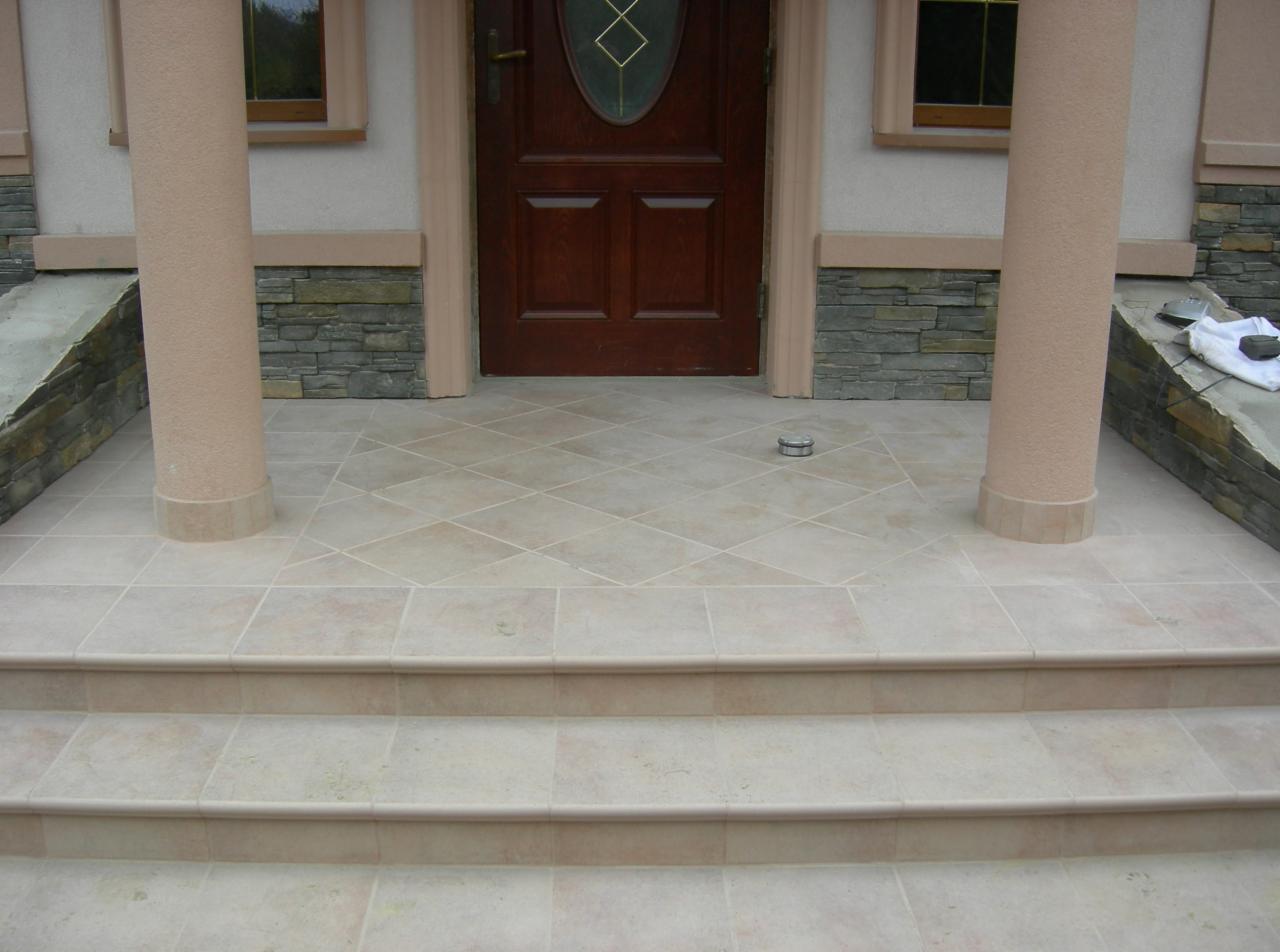 Wykończone schody wejściowe i podłoga przed drzwiami, wyłożone jasnymi płytkami ceramicznymi z wzorem rombów, kolumny w kolorze beżowym podtrzymujące zadaszenie, ciemnobrązowe drzwi wejściowe z szybą.