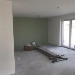 Wnętrze w trakcie remontu: ściana pomalowana na oliwkowy kolor, białe ściany, rozłożony podest malarski, karton i grzejnik na podłodze, surowa podłoga, okno balkonowe.