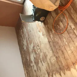 Parkiet w trakcie renowacji z widocznymi śladami starej powłoki i szlifierką do drewna, pomarańczowy kabel na podłodze, ściana pomalowana na dwa kolory.