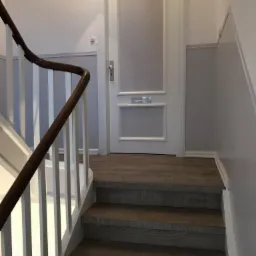 Schody wyłożone panelami podłogowymi w odcieniach szarości, z białymi balustradami i pomalowanymi na szaro ścianami, prowadzące do białych drzwi.