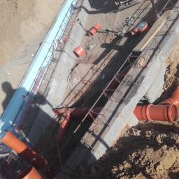 Hydroinvest - Montaż pomarańczowych rur kanalizacyjnych w betonowym wykopie zbrojonym stalą, widoczne elementy izolacji termicznej i cień osoby.