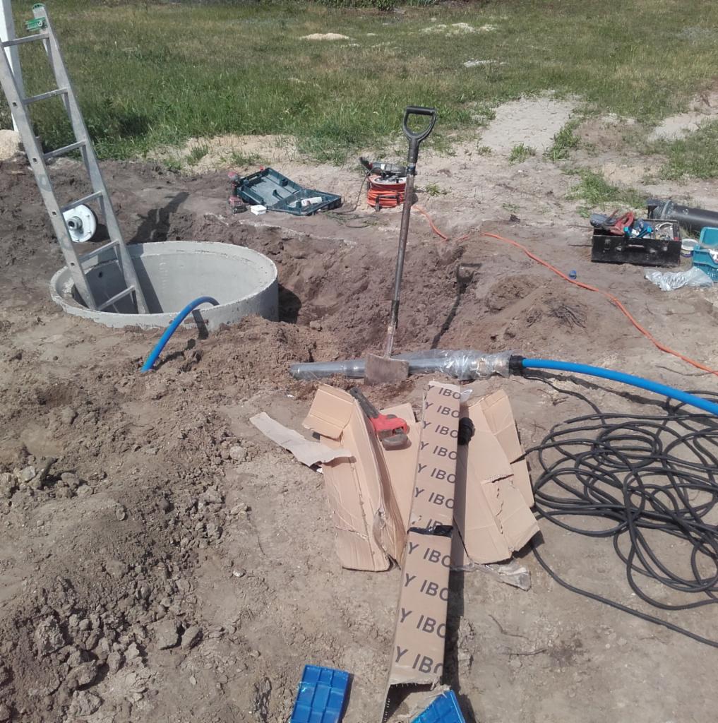 Betonowy krąg studni z drabinką w wykopie, niebieski wąż i narzędzia rozłożone na ziemi podczas prac ziemnych.