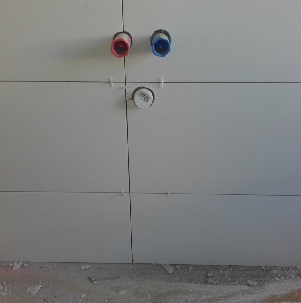 Ściana wyłożona jasnymi płytkami z widocznymi otworami na rury, oznaczone kolorami czerwonym i niebieskim, oraz otwór z zaślepką, z użyciem krzyżyków dystansowych.