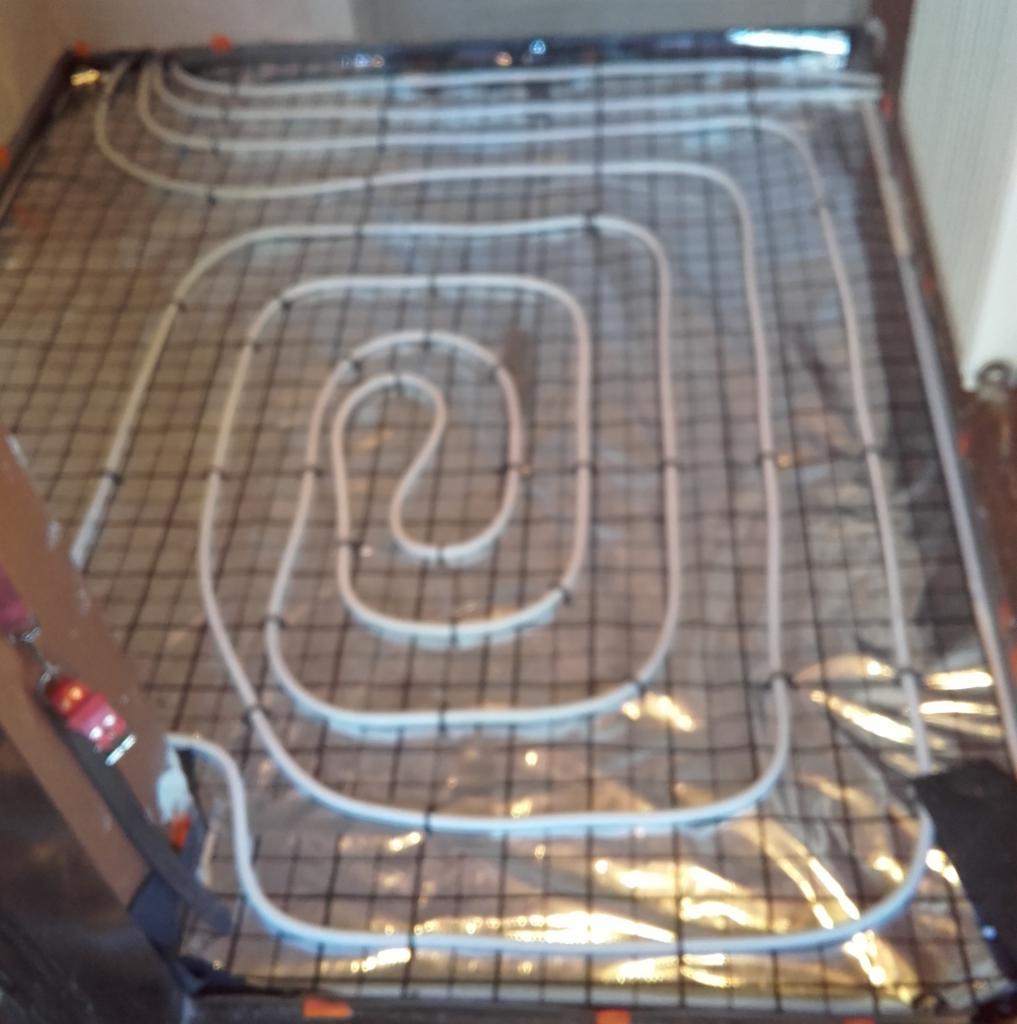 Białe rury ogrzewania podłogowego ułożone w spiralny wzór na czarnej siatce, widoczne pomarańczowe elementy mocujące na obrzeżach.