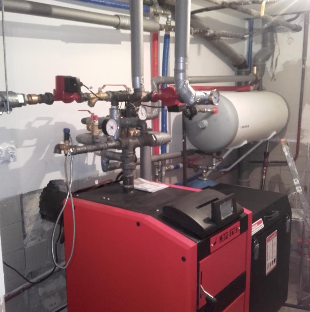 Czerwony piec CO Metal-Fach z podłączonymi rurami i manometrami w kotłowni, widoczny zasobnik ciepłej wody i drabina oparta o ścianę.