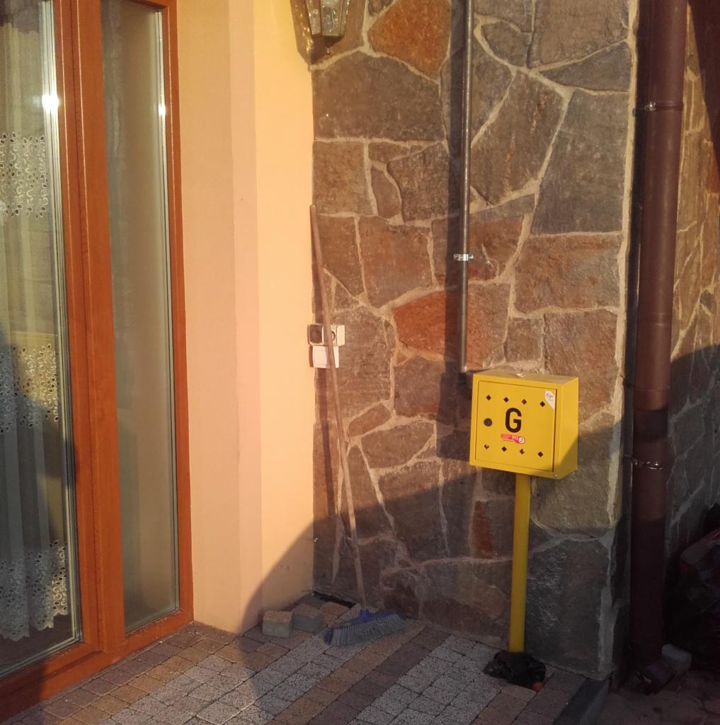 Żółta skrzynka gazowa z literą 'G' na elewacji domu wykończonej kamieniem, widoczna rura gazowa i fragment okna.