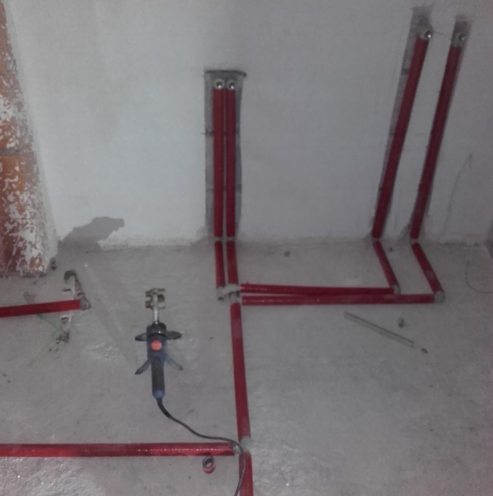 Czerwone rury instalacyjne prowadzone wzdłuż ścian w surowym pomieszczeniu, widoczny laser krzyżowy do wyznaczania poziomu, betonowa podłoga.