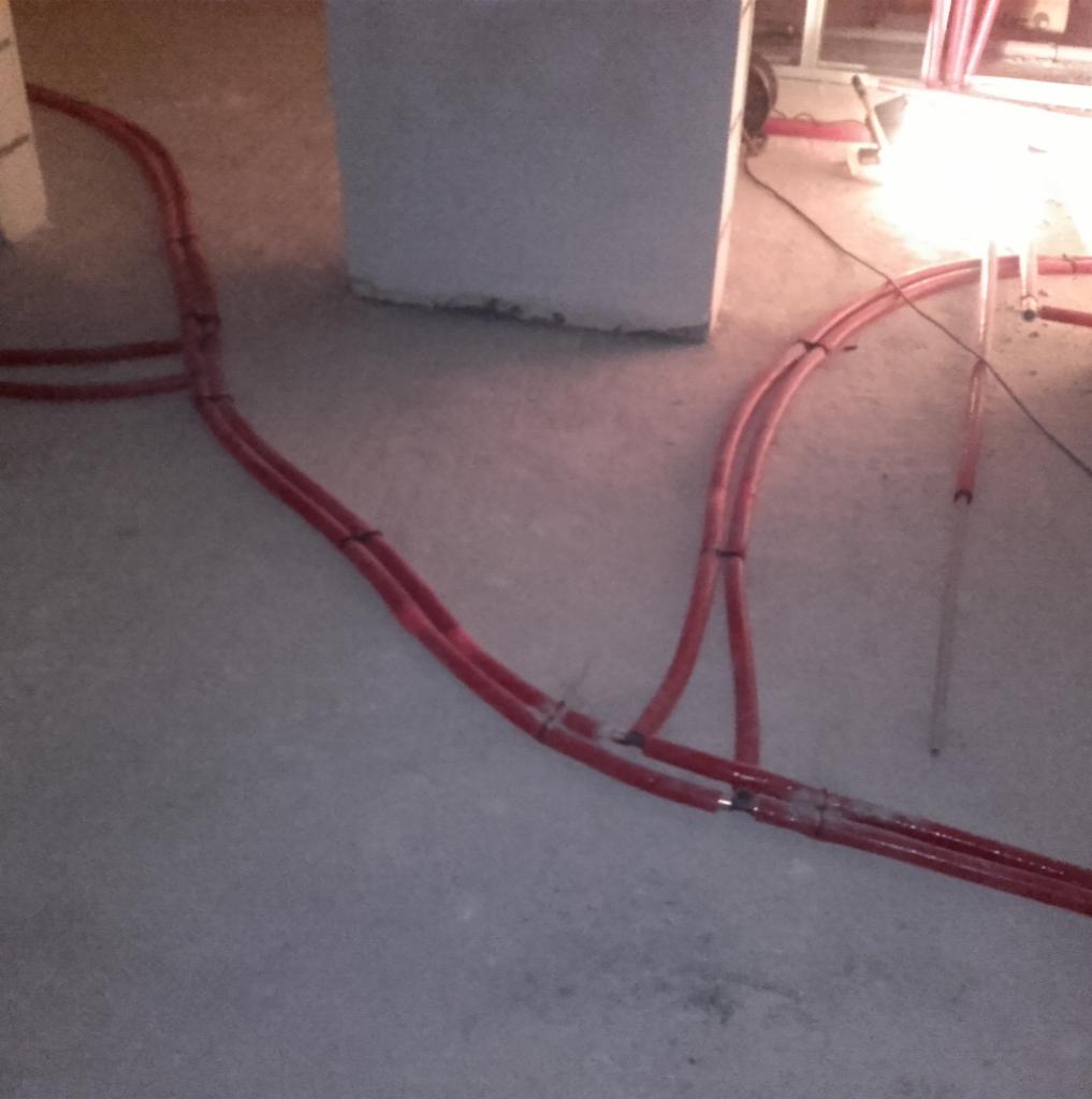 Czerwone rury instalacyjne prowadzone po szarej, betonowej podłodze w surowym wnętrzu, widoczne łączenia zabezpieczone czarną taśmą.