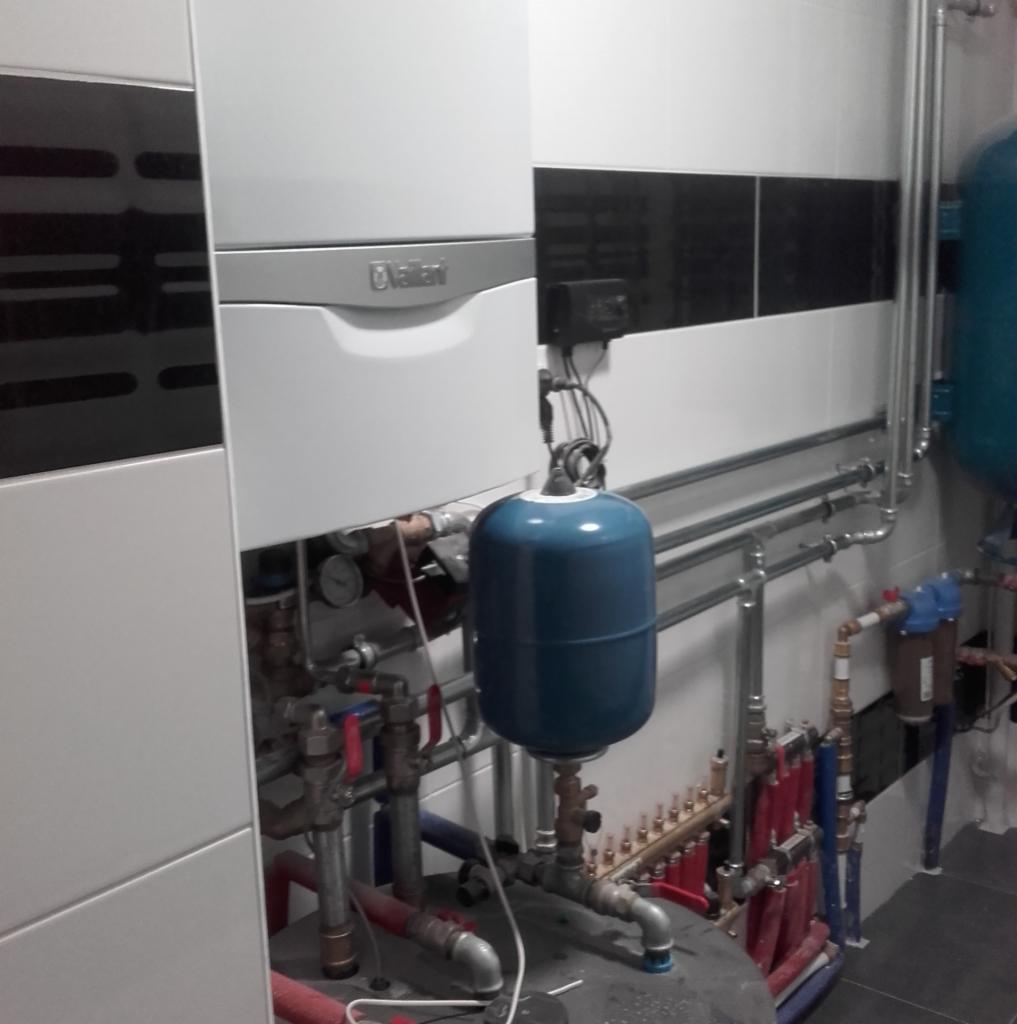 Biały piec gazowy Vaillant zamontowany na tle biało-czarnych płytek, z widocznymi rurami miedzianymi, zaworami i niebieskim naczyniem przeponowym.