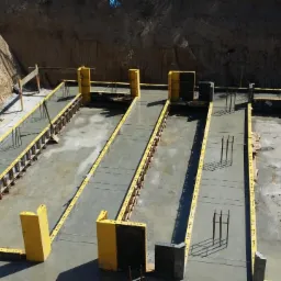 Przygotowanie do wylewania fundamentów: widoczne żółte szalunki, wystające pręty zbrojeniowe i nasypany żwir na wyrównanym terenie.