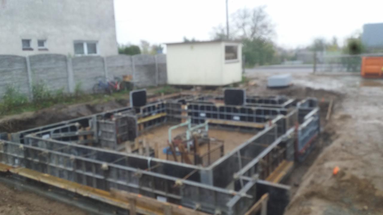 Przygotowanie szalunku pod fundament domu jednorodzinnego na działce, widoczny wykop, metalowe elementy konstrukcyjne i drewniane deski, w tle budynek gospodarczy i fragment domu.