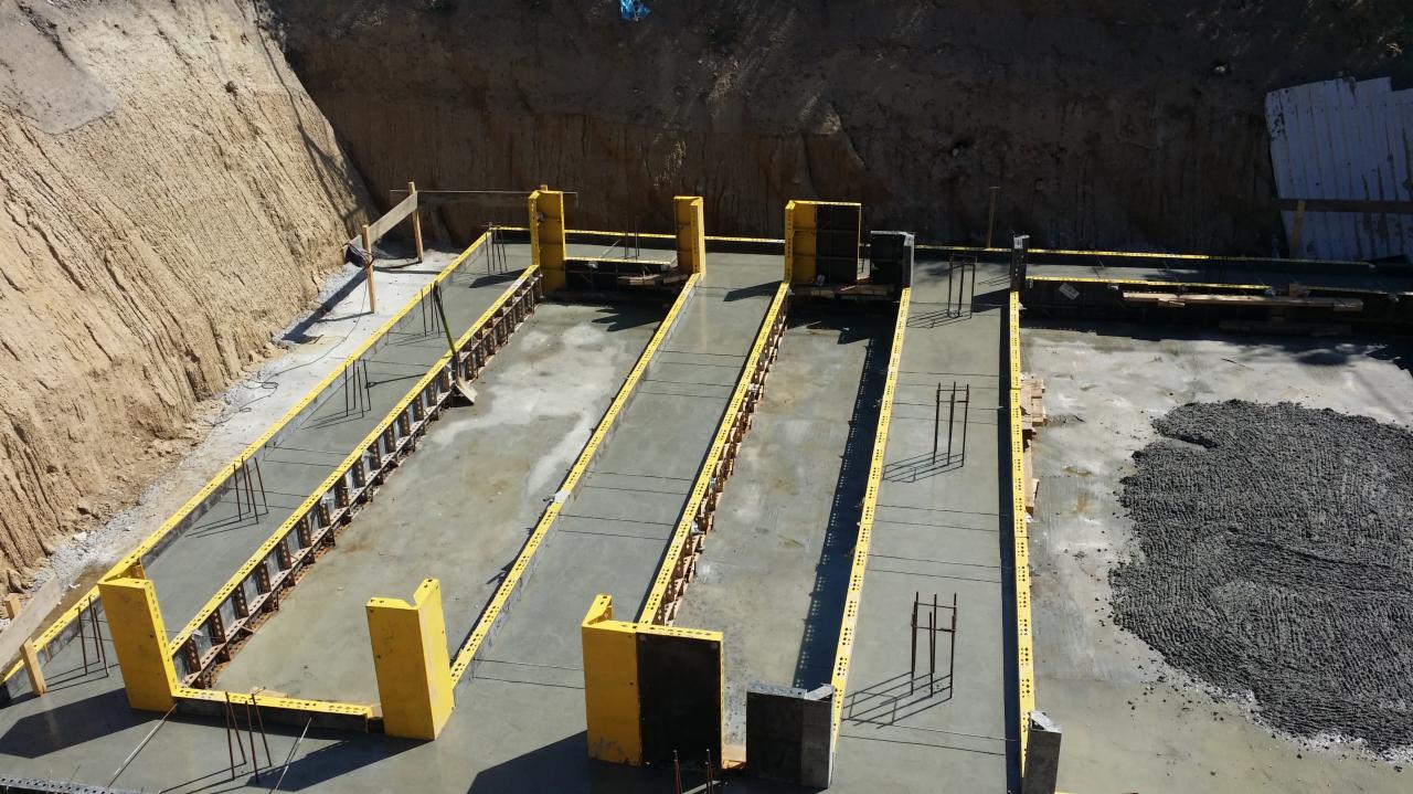 Przygotowanie do wylewania fundamentów: widoczne żółte szalunki, wystające pręty zbrojeniowe i nasypany żwir na wyrównanym terenie.