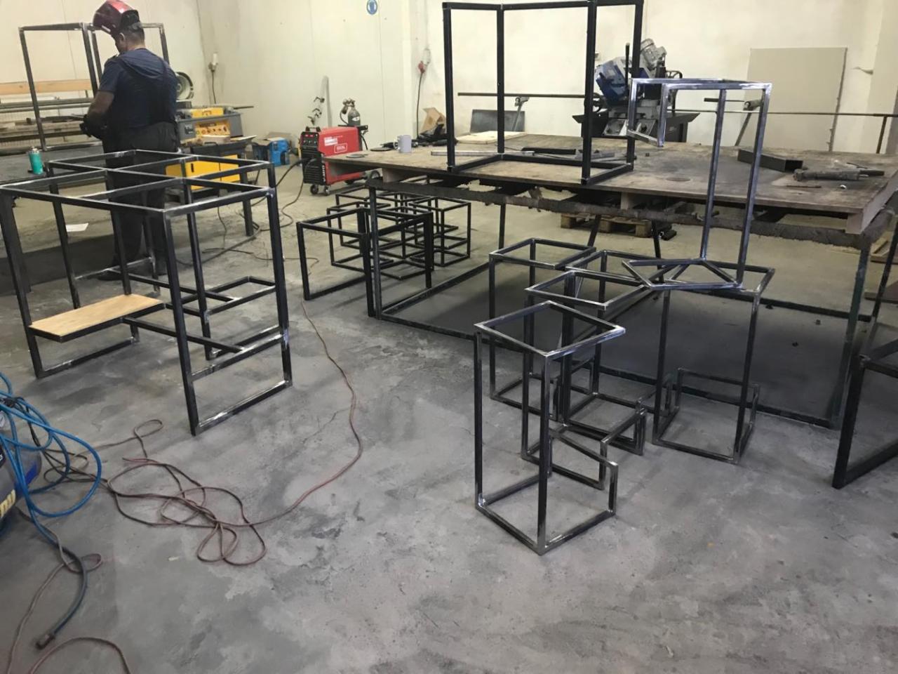 Warsztatowa sceneria z metalowymi konstrukcjami w różnej fazie wykonania, spawacz w masce ochronnej w tle, widoczne kable i narzędzia.