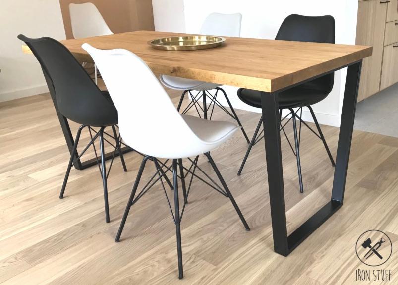 Stół z drewnianym blatem i czarnymi, metalowymi nogami w kształcie litery U, otoczony krzesłami w kolorach bieli i czerni, na drewnianej podłodze.