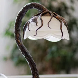 Stylowa lampka stołowa z metalową podstawą w kształcie drzewa i kloszem z białego szkła, dekorowana metalowymi liśćmi.