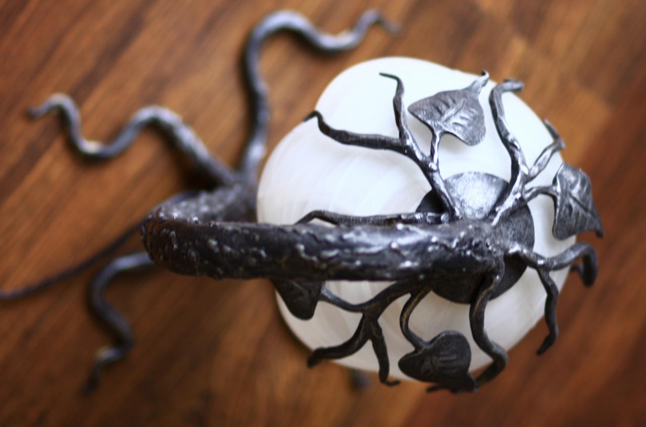 Kinkiet z kutego metalu w kształcie gałęzi z liśćmi, otaczający mleczną, okrągłą lampę na tle drewnianej ściany.