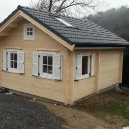 Mały, jasny domek z bali z czarnym dachem i białymi okiennicami, usytuowany na działce z widokiem na zalesione wzgórza i budynki w oddali.