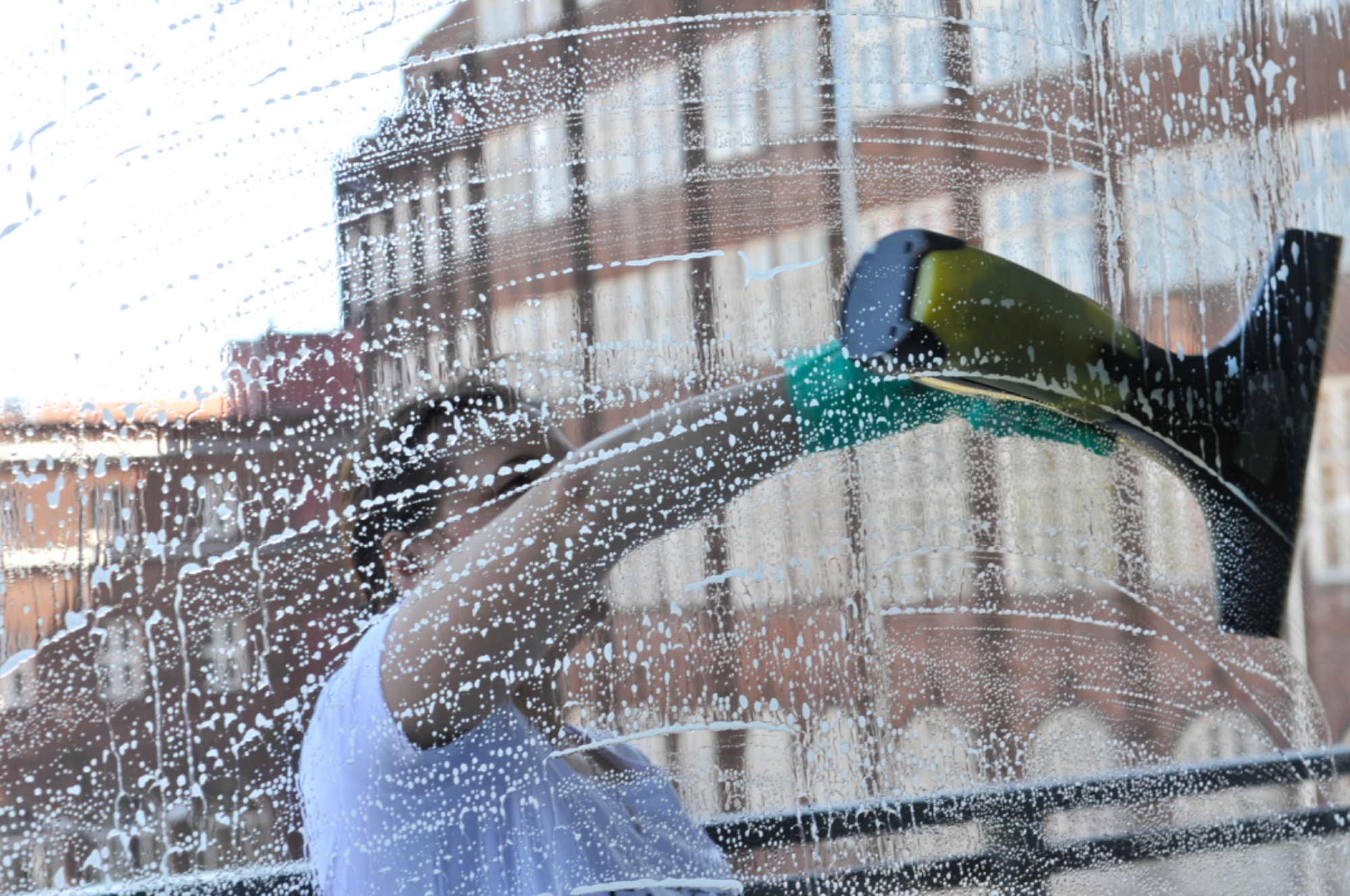 Osoba myje okno na wysokości, używając ściągaczki z zieloną gąbką; widoczny budynek w tle przez zapienione szkło.