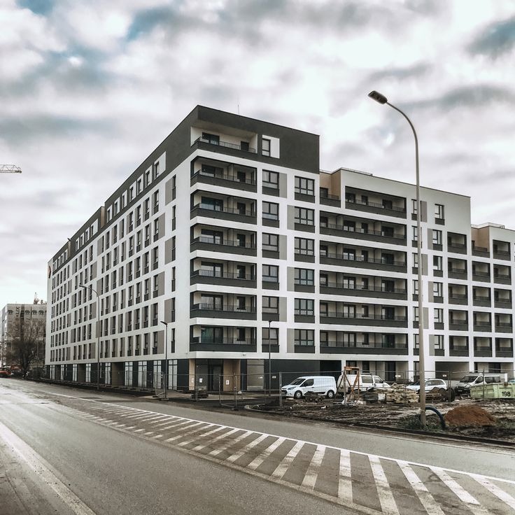 Nowoczesny, wielopiętrowy budynek mieszkalny z balkonami, widoczny z ulicy. W tle pochmurne niebo. Na pierwszym planie jezdnia z pasami. Uwagę przykuwa biała fasada budynku.